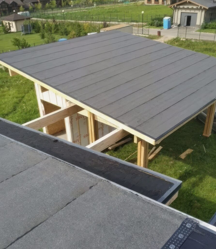 O imagine a unui acoperiș din lemn, cu o suprafață neagră, situat deasupra unei structuri în construcție, cu iarbă verde în jur.
