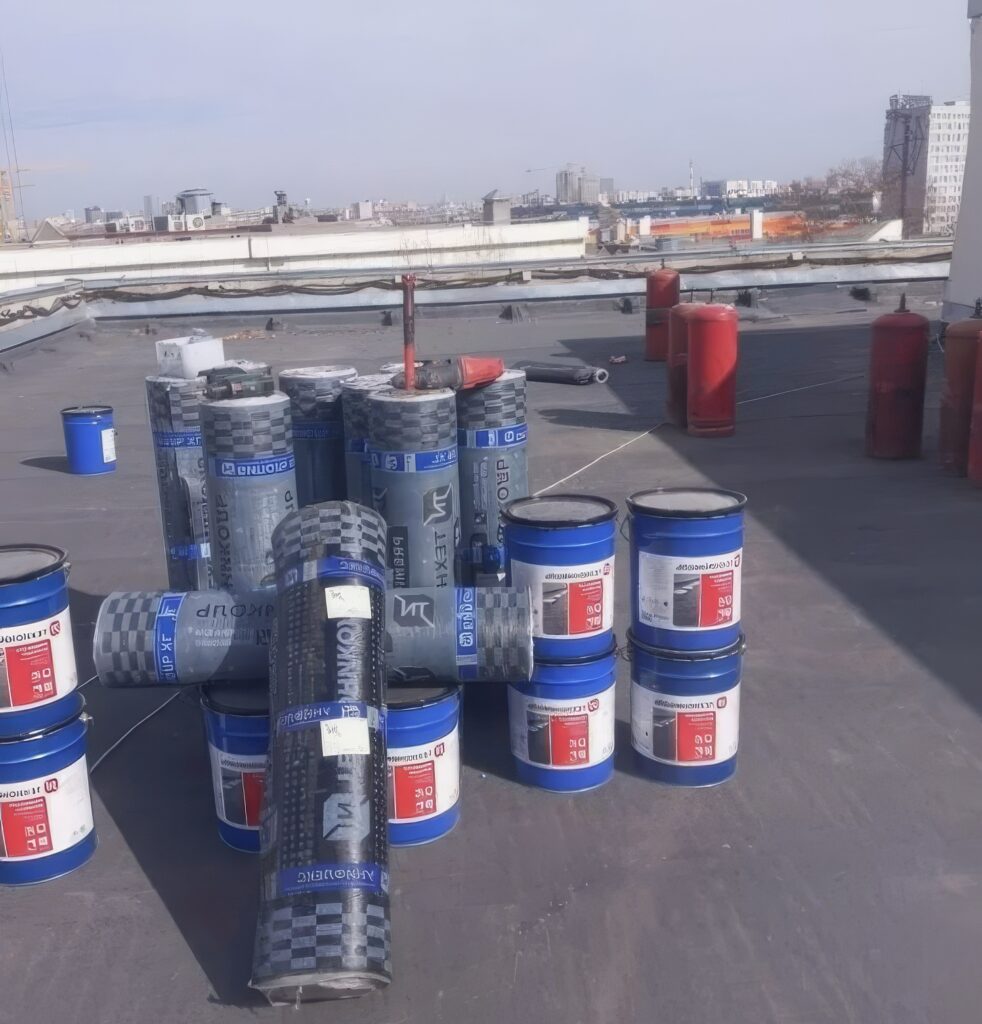 Diverse bidonuri de materiale de construcție, aranjate pe un acoperiș, cu orașul în fundal.